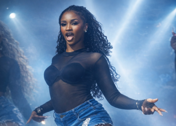 Favour “Dat Black Chi” Odimuko performing a high-energy dance routine on stage under dramatic blue lighting, wearing a black performance top, denim fringe shorts, and white leg warmers.
