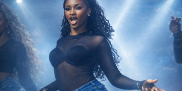 Favour “Dat Black Chi” Odimuko performing a high-energy dance routine on stage under dramatic blue lighting, wearing a black performance top, denim fringe shorts, and white leg warmers.